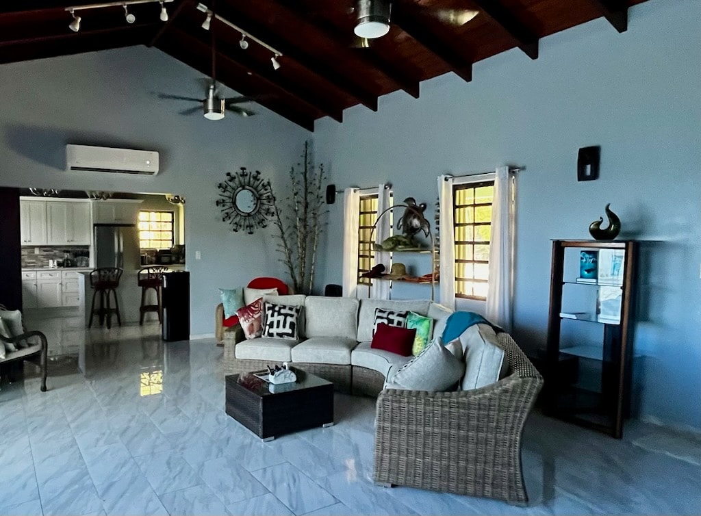 A spacious great room features high ceilings with exposed wooden beams and large windows allowing natural light to brighten the area. Comfortable seating is arranged around a central coffee table, with decorative accents visible on shelves and walls.