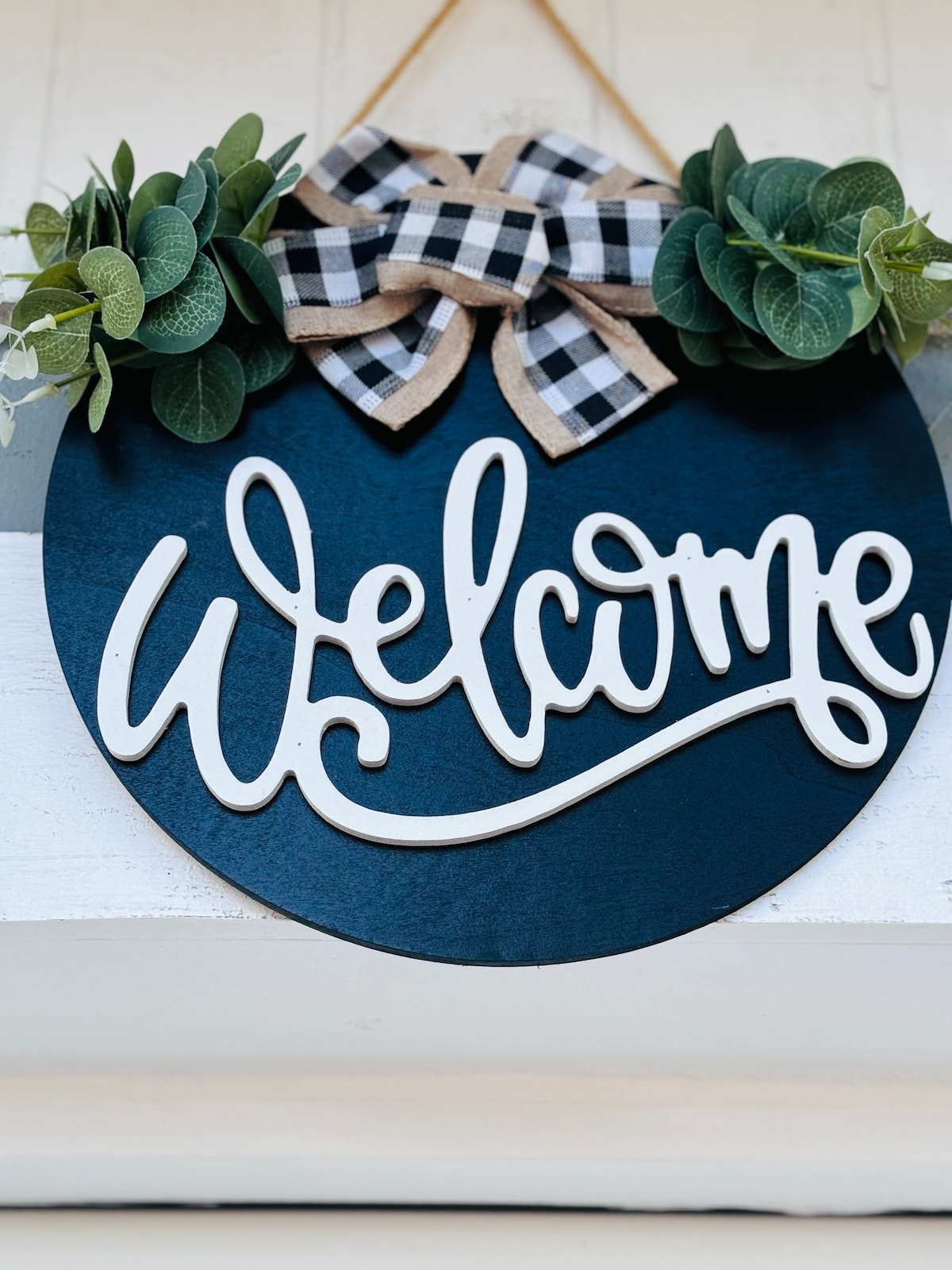 A circular sign featuring the word 'Welcome' in white, crafted with a decorative cursive font. The sign is accented with a black and white checkered bow and adorned with greenery, providing an inviting entryway touch.