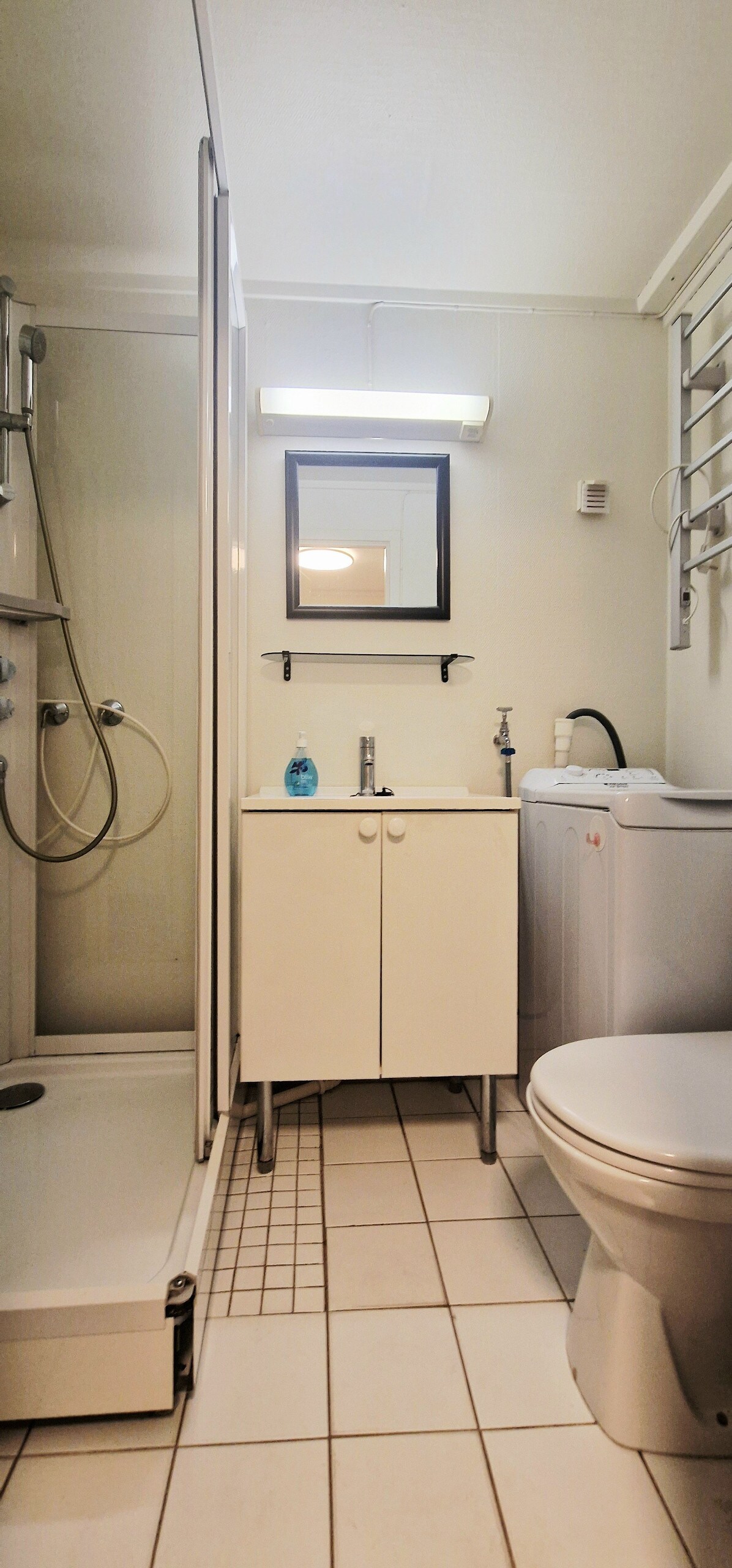A modern bathroom is equipped with a shower stall and a compact vanity with storage. A mirror hangs above the sink, and a washing machine is positioned beside the door. Neutral-toned tiles cover the floor, and bright lighting illuminates the space.