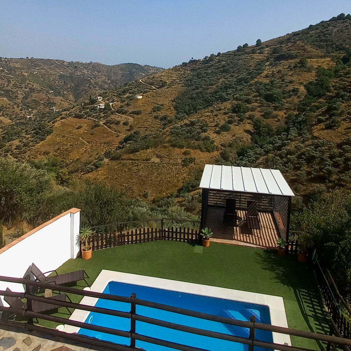 The outdoor area features a private pool surrounded by grass and potted plants. A shaded seating area is visible near the pool. The expansive view showcases rolling hills and valleys, highlighting the natural beauty of the Andalusian countryside.