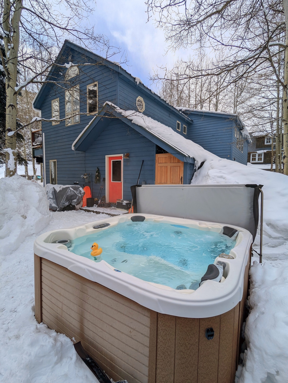 A private hot tub is situated in a snowy outdoor area, with clear blue water visible. The blue exterior of the two-story house is depicted behind the hot tub, blending into a serene mountain setting. Snow drapes over the ground and surrounding structures.
