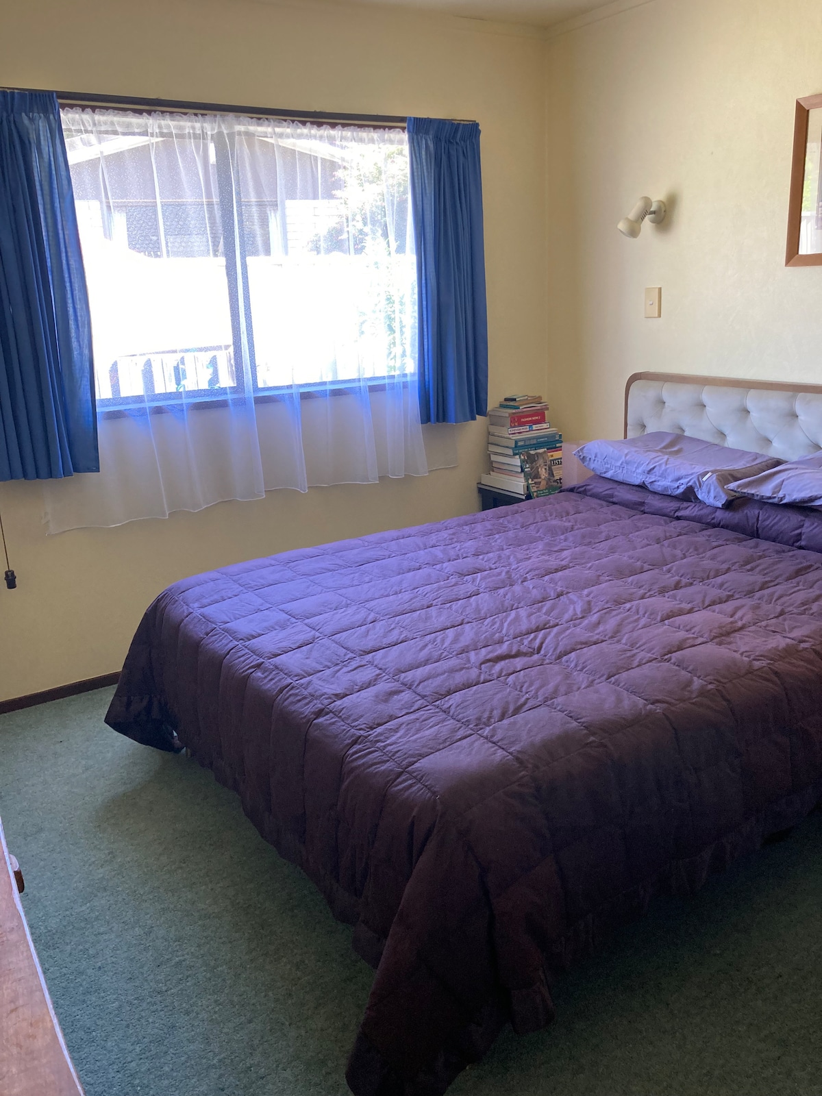 A cozy bedroom features a spacious bed covered with a purple quilt. Natural light filters through a window adorned with sheer curtains. A small stack of books is organized by the window, complementing the overall inviting ambiance of the room.