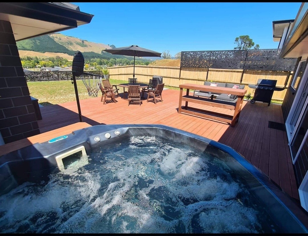 A large hot spa is positioned on the deck, with bubbling water visible. Outdoor furniture, including several wooden chairs and a table, is arranged in the background under a sun umbrella. A grassy area and scenic hills can be seen beyond the deck.