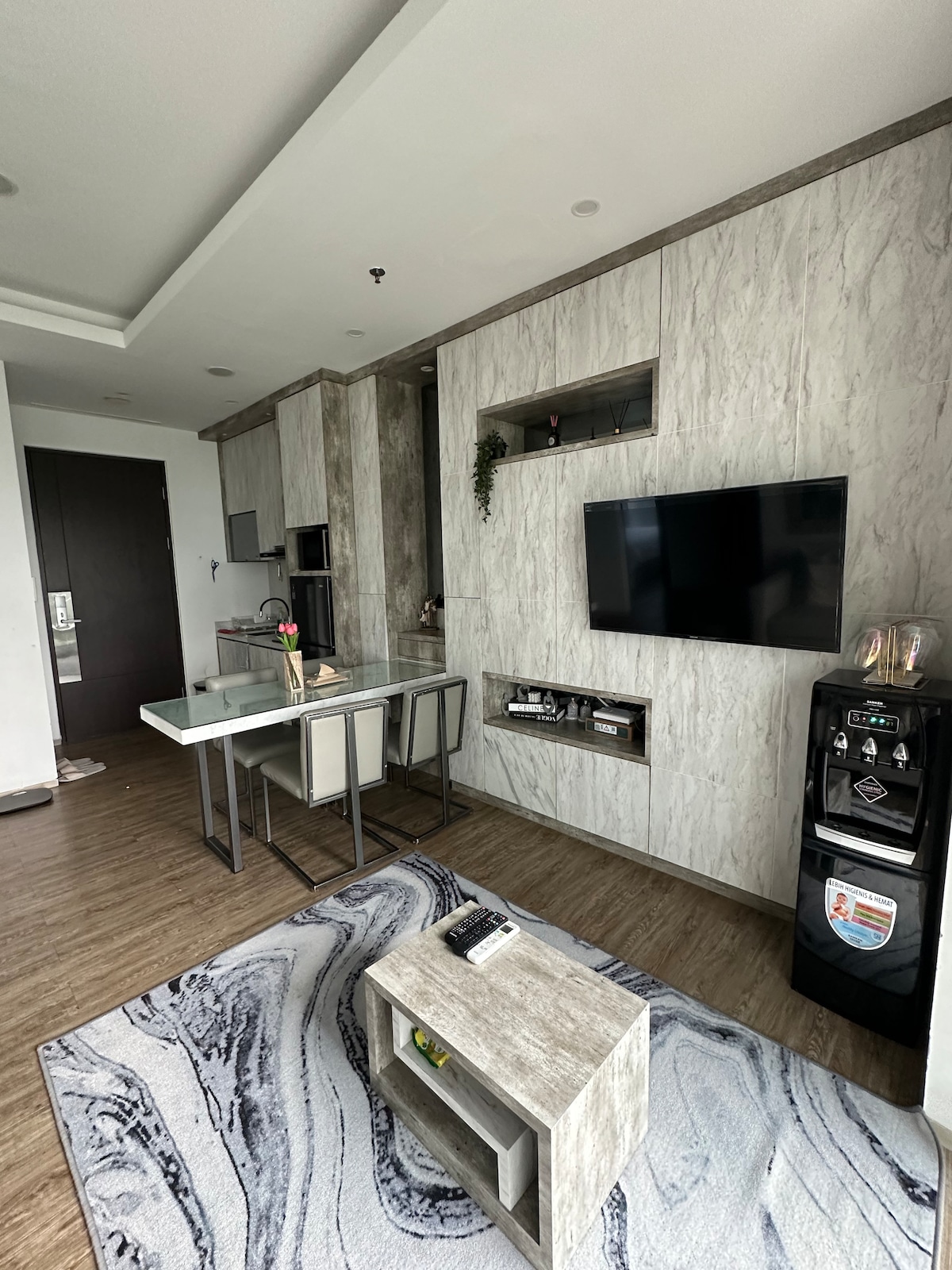 A modern living area features a light-colored table with four chairs, positioned near a wall-mounted television. A sleek cabinet provides storage under the TV. Distinctive marble-patterned carpet adds texture to the space, complemented by an elegant, minimalist design throughout.