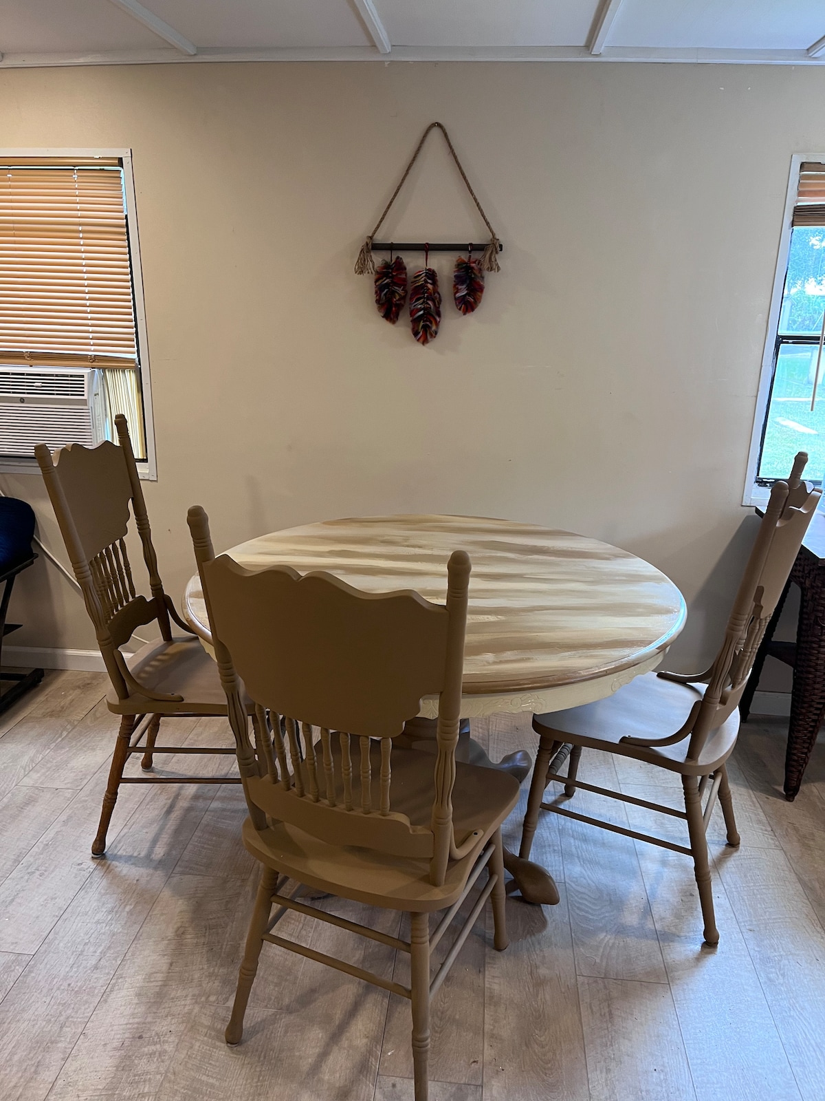 A round dining table is set in a well-lit corner, surrounded by four wooden chairs. Natural light streams through the nearby window, highlighting the simplicity of the decor. A wall hanging featuring feathers adds a touch of character to the space.