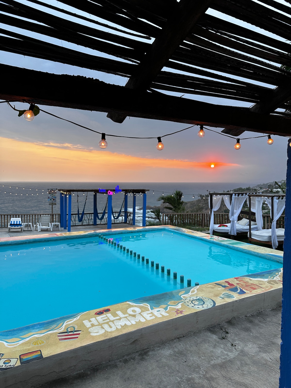 A vibrant swimming pool is framed by a wooden pergola, adorned with string lights that create a relaxed atmosphere. The sunset casts a warm glow over the water, while the horizon reveals the silhouette of the ocean. Lounge chairs are arranged for comfort nearby.