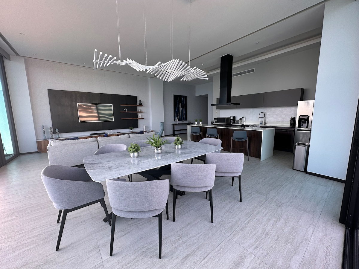 A modern dining area features a large marble table surrounded by six chairs, complemented by a stylish light fixture above. The open space leads to a sleek kitchen equipped with stainless-steel appliances, seamlessly merging dining and cooking areas with large windows letting in natural light.