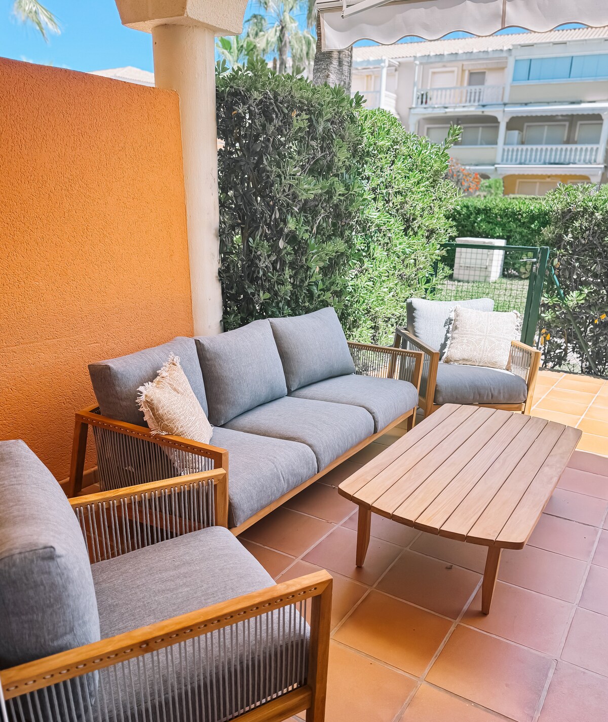 A cozy outdoor terrace is featured, furnished with a wooden coffee table and a grey cushioned seating area, including a pair of armchairs. Lush green foliage surrounds the space, providing privacy and a sense of tranquility. A sunshade offers protection from direct sunlight.