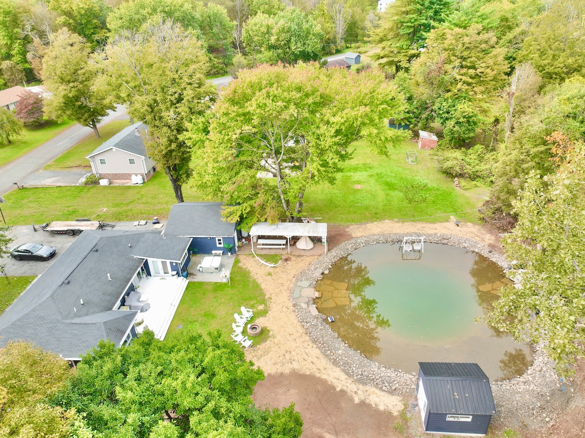 An aerial view captures the property layout, featuring a swim pond surrounded by gravel and natural landscaping. A picnic area with seating is visible, along with a tree shading the outdoor space. Nearby, the house is complemented by lush greenery and residential streets beyond.