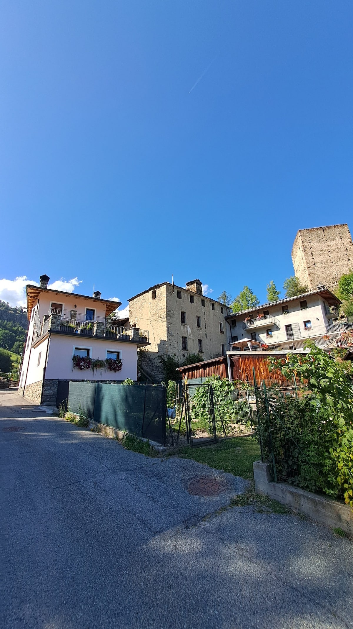 A quiet street in Gignod is depicted, featuring a mix of stone and wooden structures. The clear blue sky overhead contrasts with the greenery along the roadside. A charming blend of buildings provides a glimpse of the local architecture set against a mountainous backdrop.
