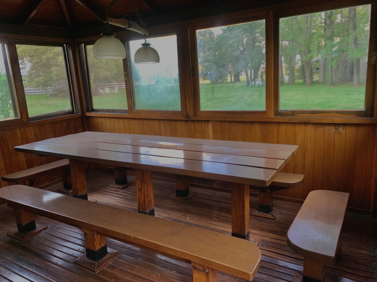A spacious dining area is showcased with a long wooden table and benches, all set against a backdrop of large windows that offer views of the green surroundings. Natural light fills the space, complemented by two hanging light fixtures overhead.