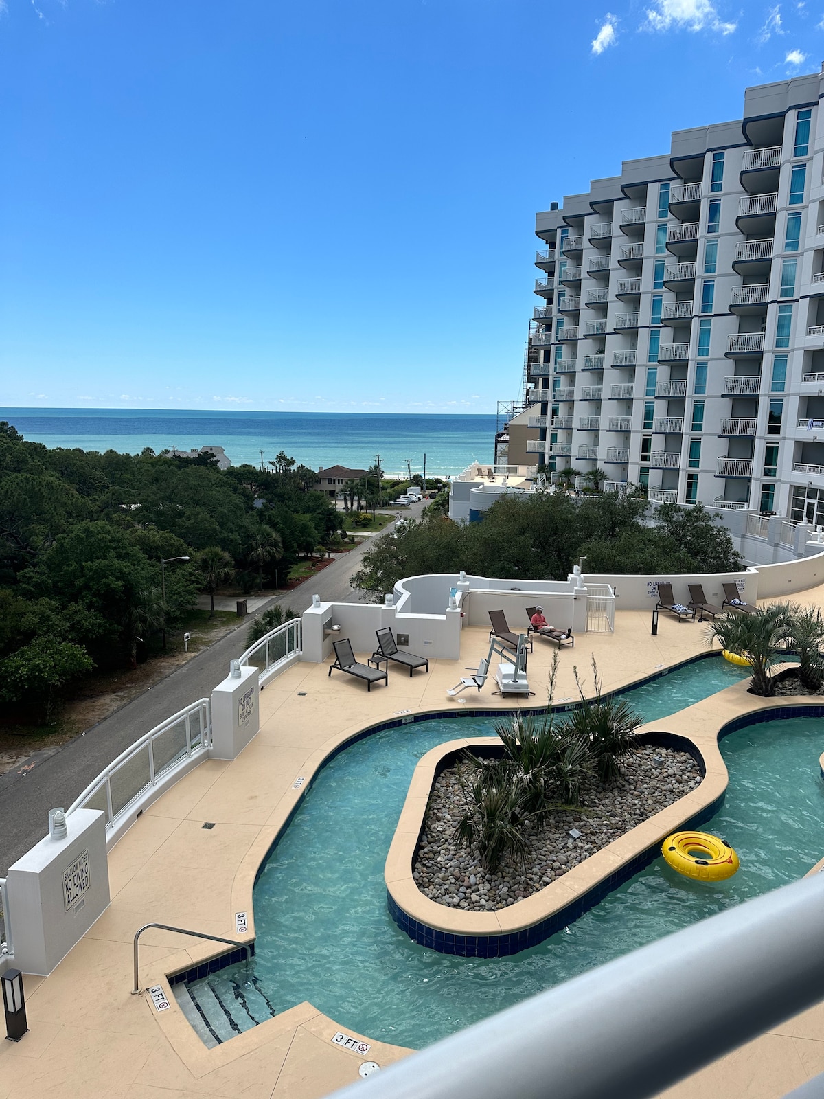 The view from the balcony captures a sunny outdoor pool area winding through lush greenery, leading towards the ocean. The sparkling water is visible in the background, along with a clear blue sky. Lounge chairs are arranged around the pool, inviting relaxation.