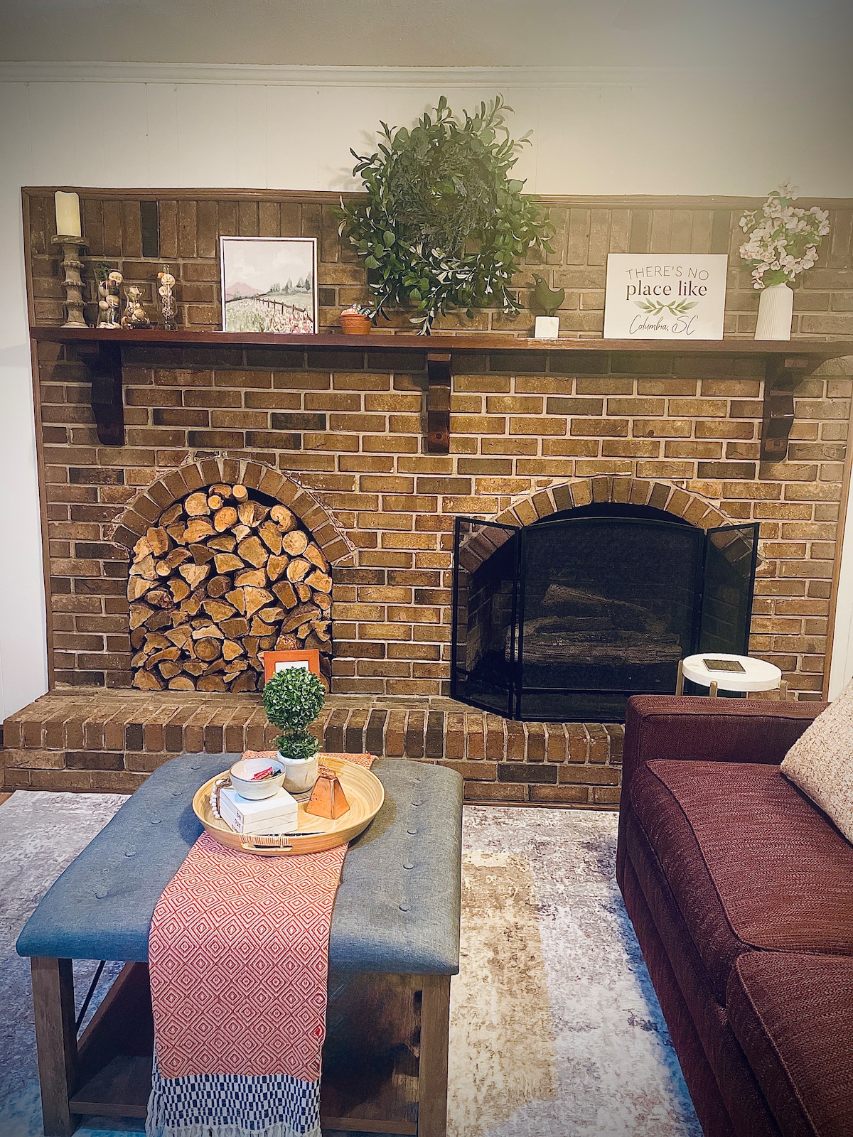 A cozy living area features a brick fireplace with a neatly stacked wood log pile on the left. Above the fireplace, decorative items and a framed picture are displayed. A coffee table with a tray holding an arrangement of items sits in front of a comfortable brown sofa.