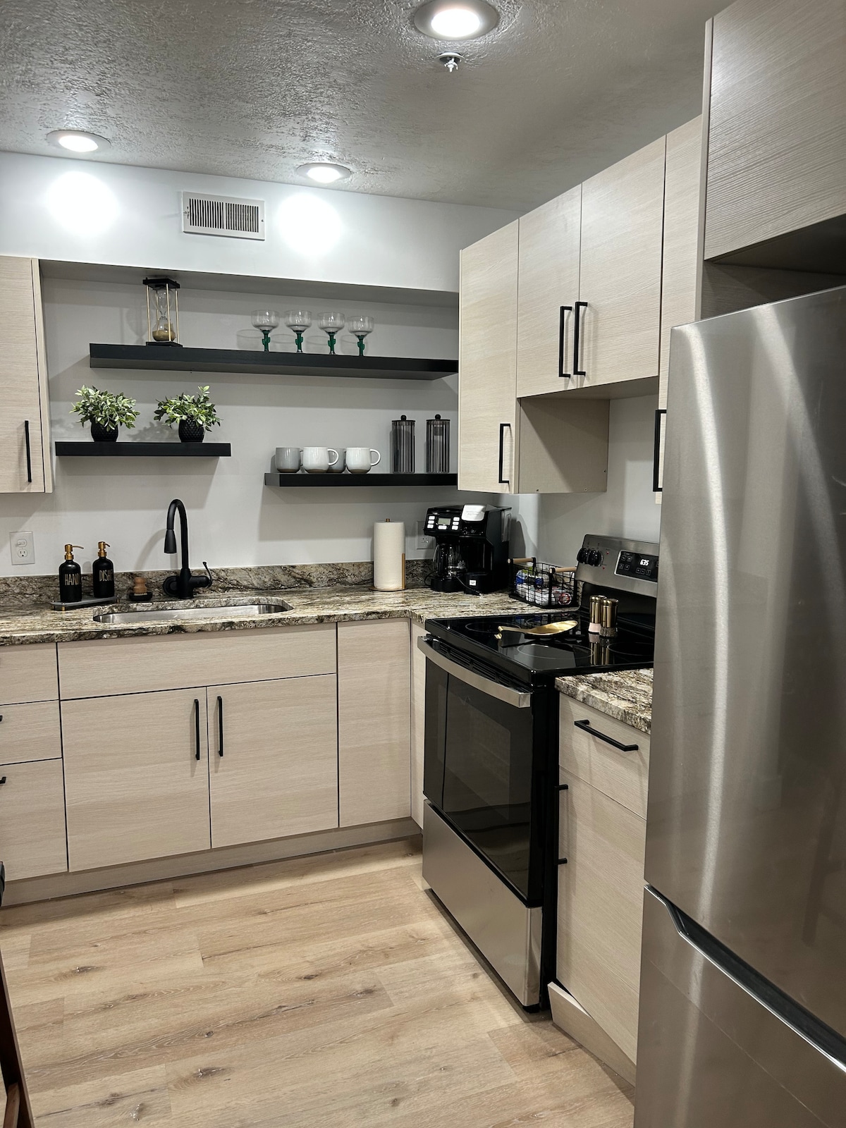 A modern kitchen is equipped with stainless steel appliances and a granite countertop. Open shelving displays glassware and coffee essentials. Neutral cabinetry complements the design, while under-cabinet lighting enhances functionality. A black sink and faucet are positioned between the stove and refrigerator, creating an efficient layout.