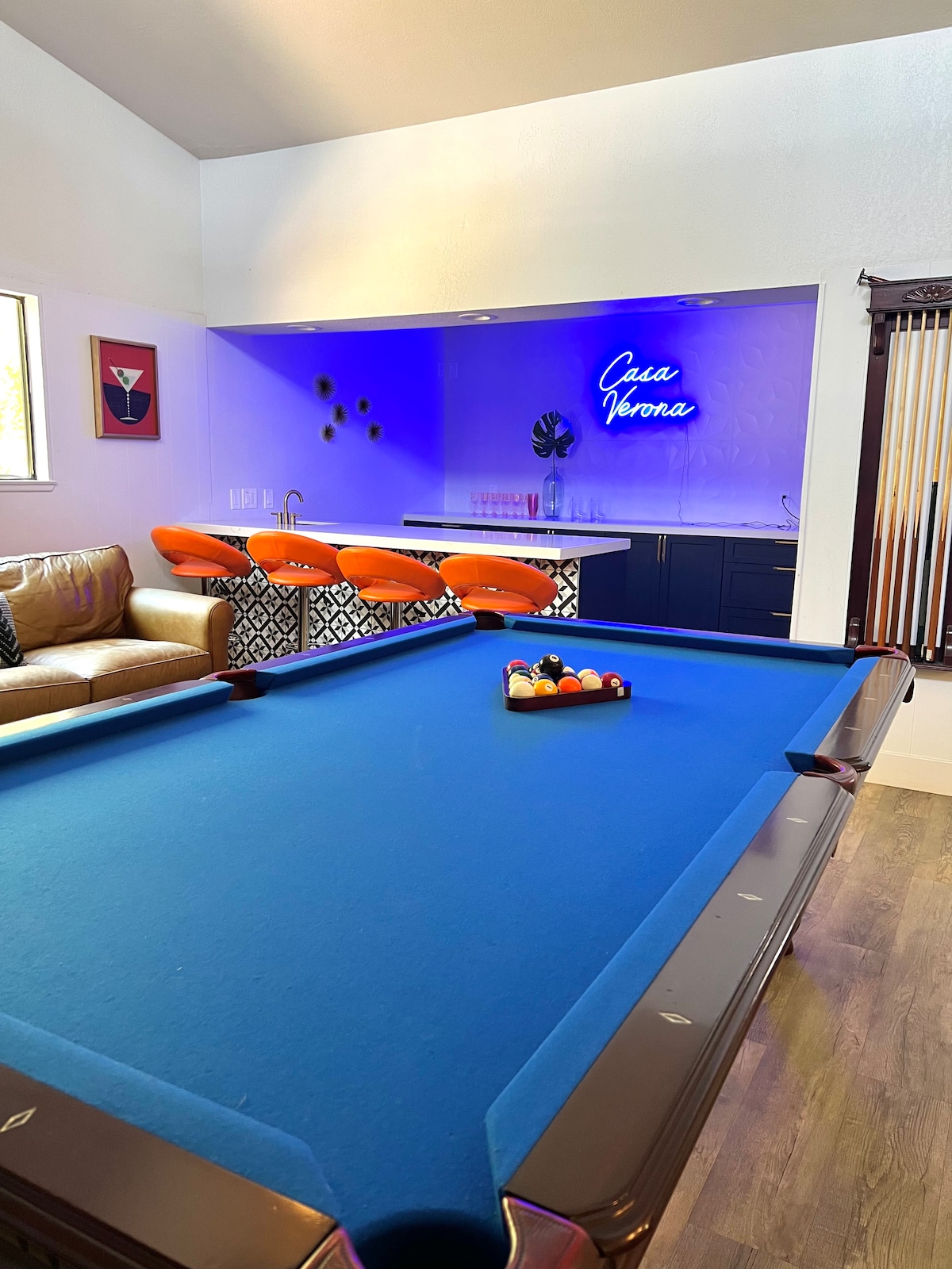 A game room is featured, showcasing a blue felt pool table with a set of colored balls arranged on it. A cozy seating area with a tan couch is visible, complemented by vibrant orange bar stools against a stylish black and white patterned bar. Soft lighting enhances the ambiance.