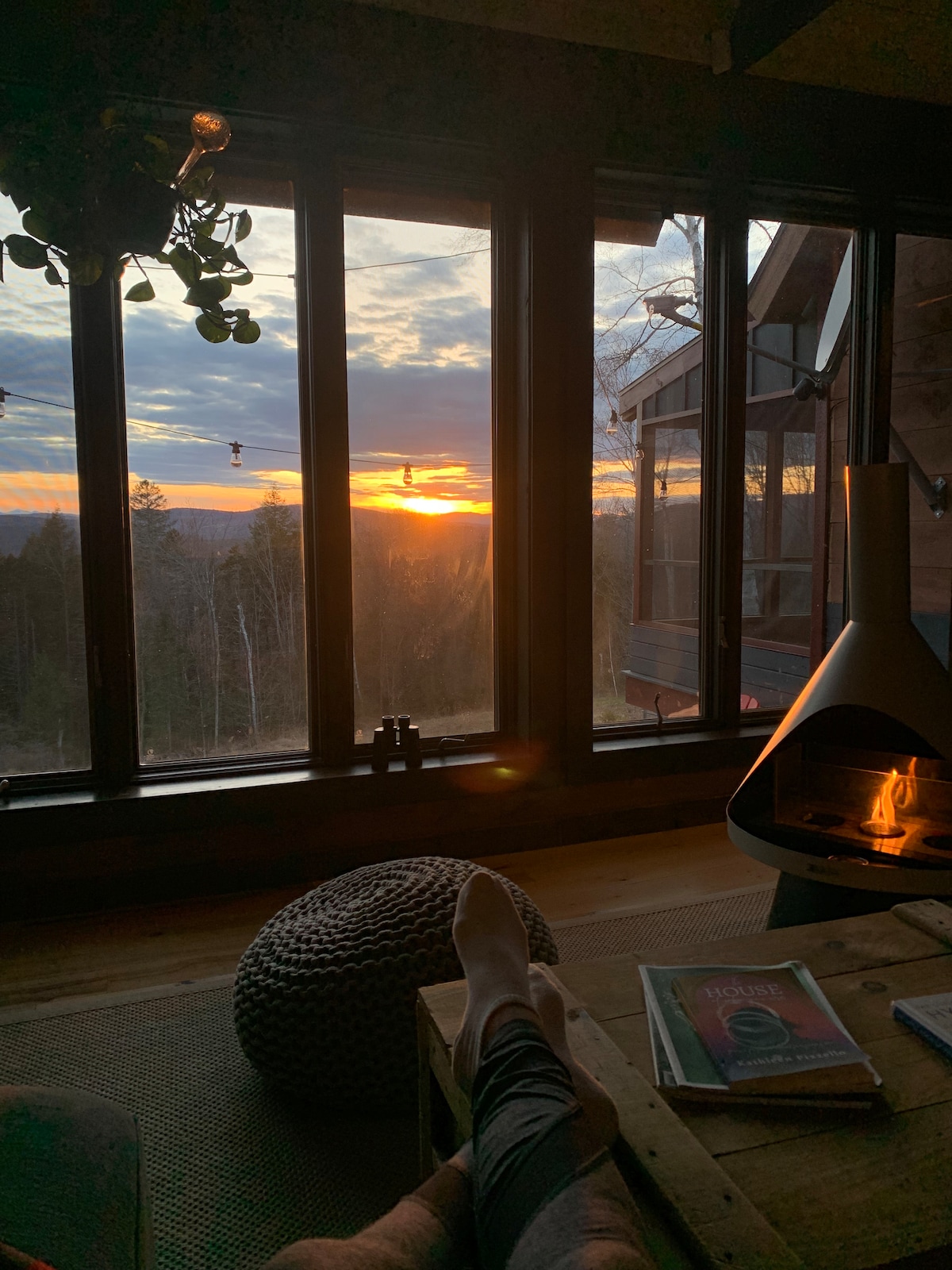 A cozy corner is captured with a view of the sunset through large windows. A softly glowing fire burns in a modern fireplace, and a magazine rests on a wooden coffee table. A calming atmosphere is enhanced by natural light and outdoor scenery.