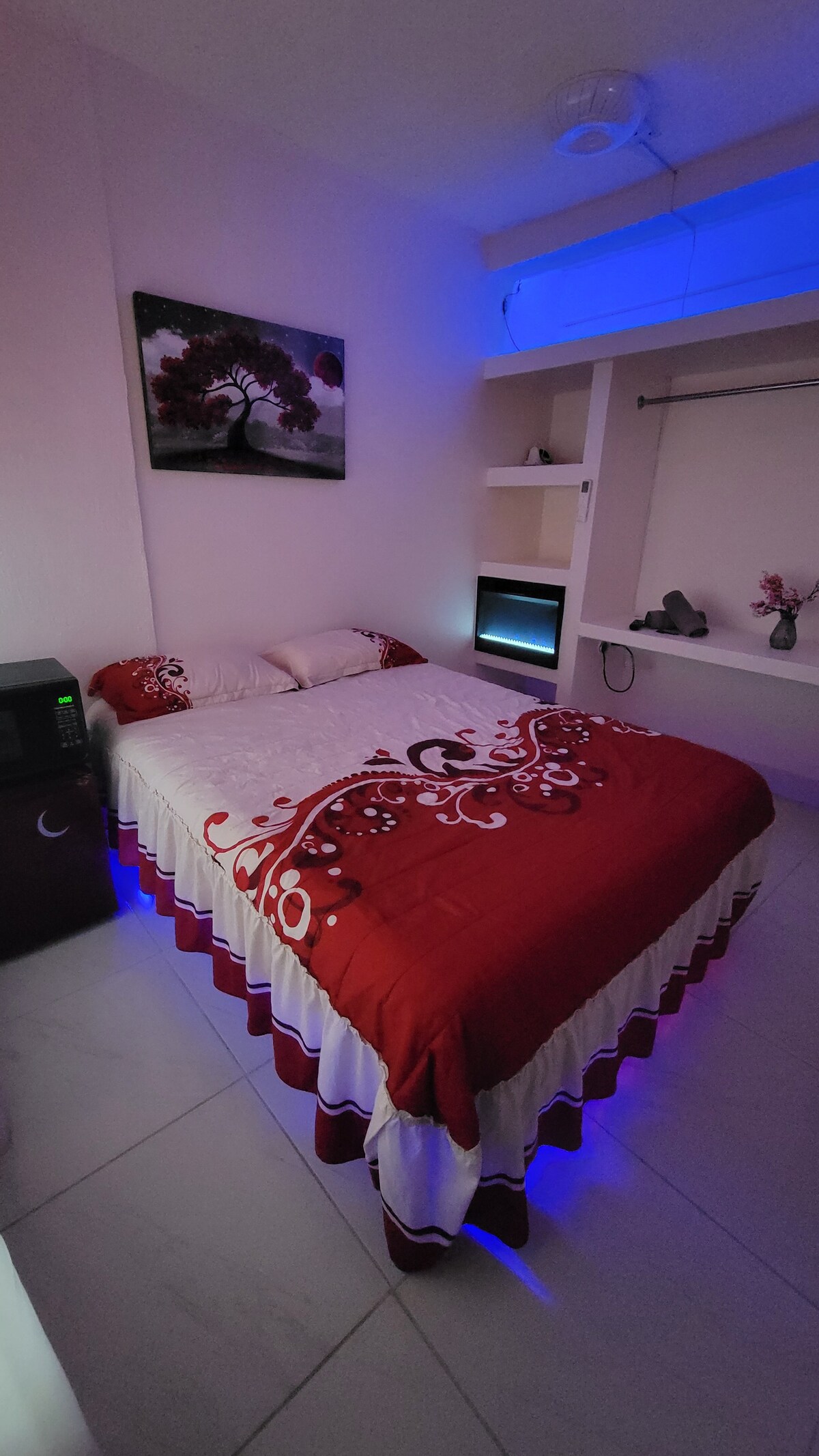 A cozy sleeping area is presented with a spacious bed dressed in a red and white comforter. Soft blue lighting enhances the ambiance. A small refrigerator and a TV are positioned within the room, while a shelf holds decorative items and additional storage.