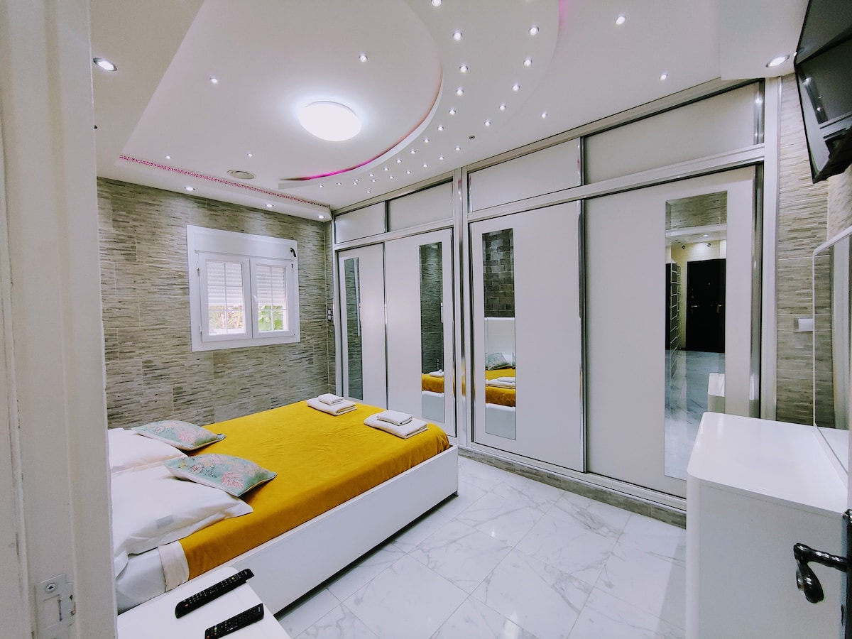 A bright bedroom is presented with a spacious bed featuring an orange and white bedding set. Sleek mirrored wardrobes reflect the room's light, and modern recessed lighting illuminates the ceiling. A white dresser stands beside the bed, complementing the room's contemporary design.