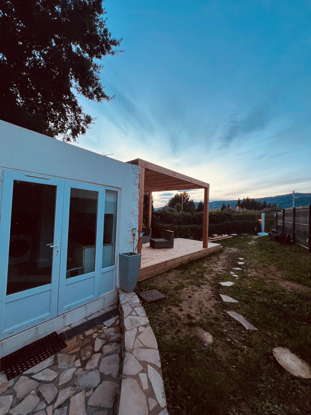 A modern exterior view reveals a terrace with a wooden overhang, designed for relaxation. Stone pathways lead from the entrance to the lush garden, while the soft glow of the evening sky and surrounding greenery enhance the tranquil setting.