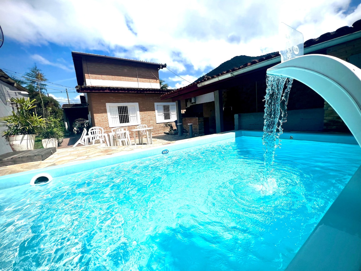 A swimming pool is featured with a gentle waterfall cascading into the clear blue water. Sunlight reflects on the surface, creating a serene atmosphere. Surrounding the pool, a patio area is equipped with several white outdoor chairs and a table, ideal for relaxation.