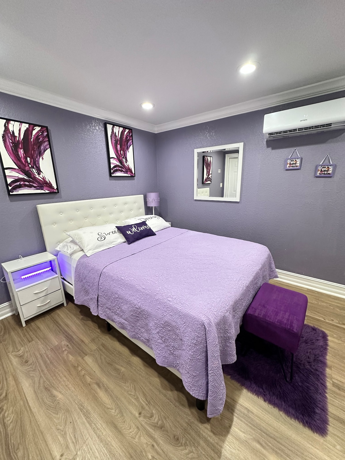A serene sleeping area features a spacious bed dressed in purple linens, complemented by two decorative wall art pieces in shades of purple. A nightstand with a soft-lit lamp sits beside the bed, and a small stool provides additional seating, enhancing the room's inviting atmosphere.
