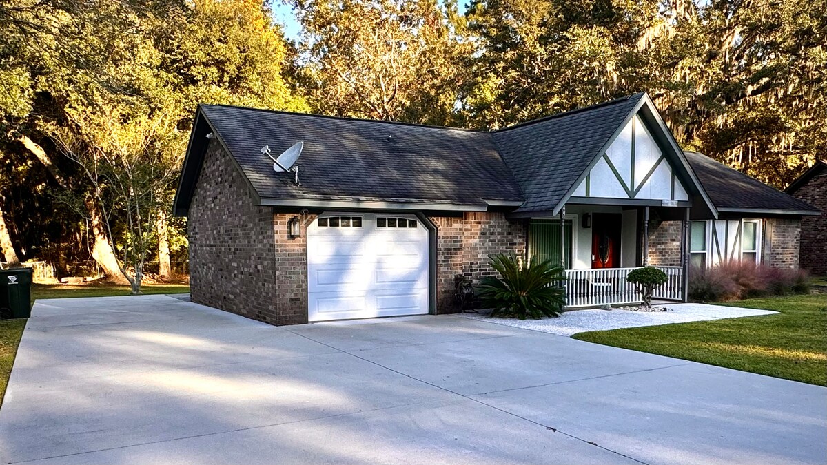 A ranch-style house composed of brick features a sloped roof and a one-car garage. A manicured lawn surrounds the home, with shrubs and trees in the background. The front porch is framed by white columns, providing an inviting entryway.