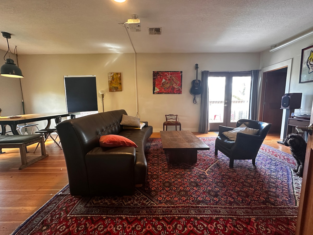 An open living and dining area is furnished with a dark leather sofa and a wooden coffee table atop a patterned rug. A dining table is situated in the background, with a large window offering access to a deck. Nearby, a comfortable chair invites relaxation.