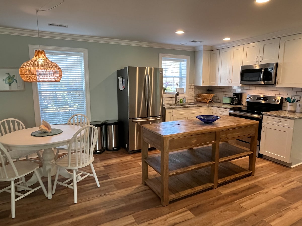 The kitchen features light cabinetry, a spacious wooden island, and modern stainless steel appliances. A round dining table with white chairs is positioned beside a window, allowing natural light. Soft green walls provide a gentle backdrop, creating a welcoming atmosphere for dining or cooking.