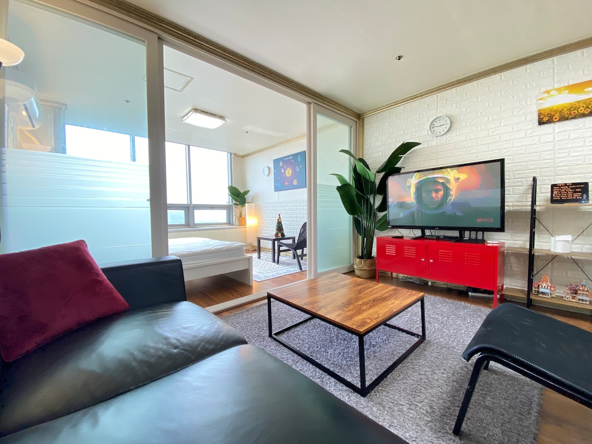 The living area features a comfortable black sofa alongside a coffee table set on a plush rug. A television is mounted on a stand with decorative items. A glass partition separates the sleeping area, with a bed visible in the background, illuminated by natural light.