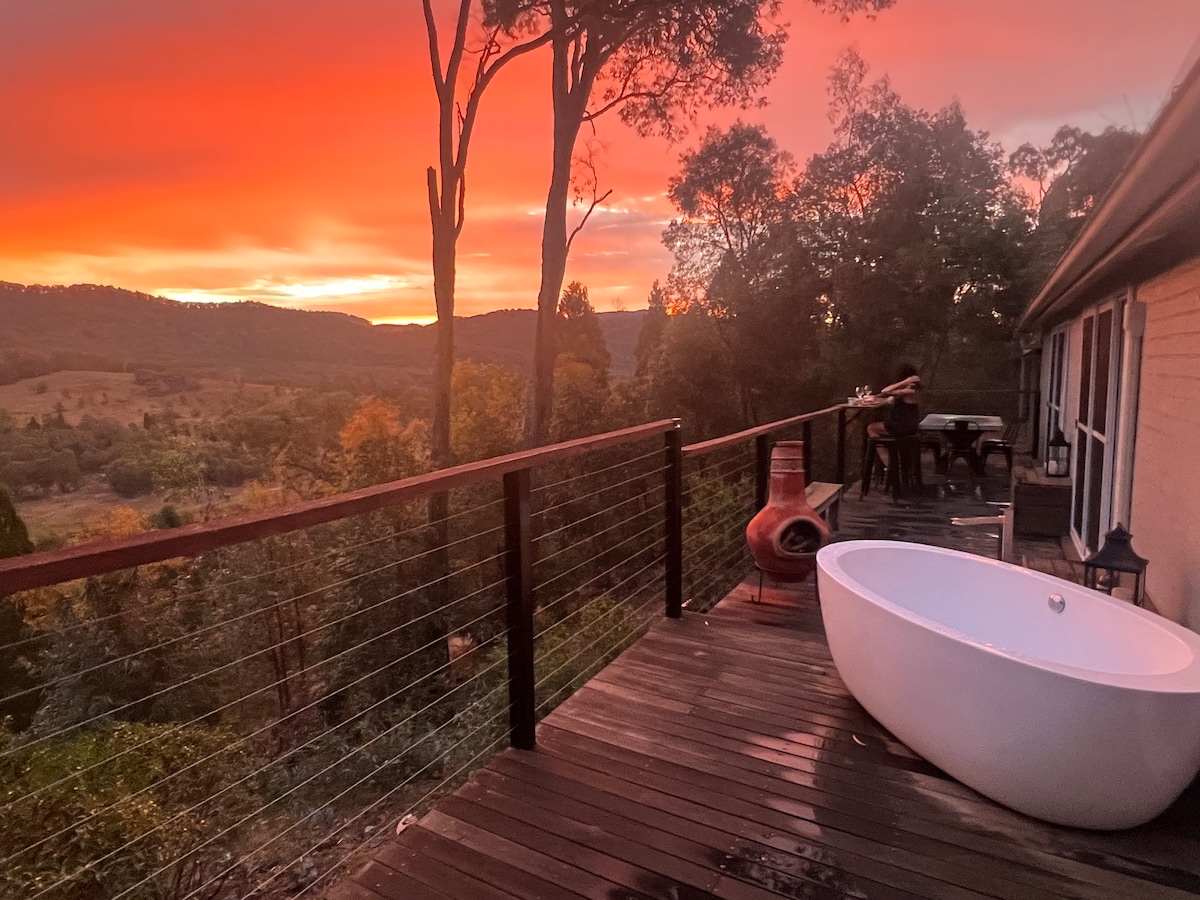An outdoor deck is revealed, featuring a freestanding bathtub and a seating area. The evening sky is painted in vibrant shades of orange and pink, casting a warm glow over the surrounding natural landscape of hills and trees.