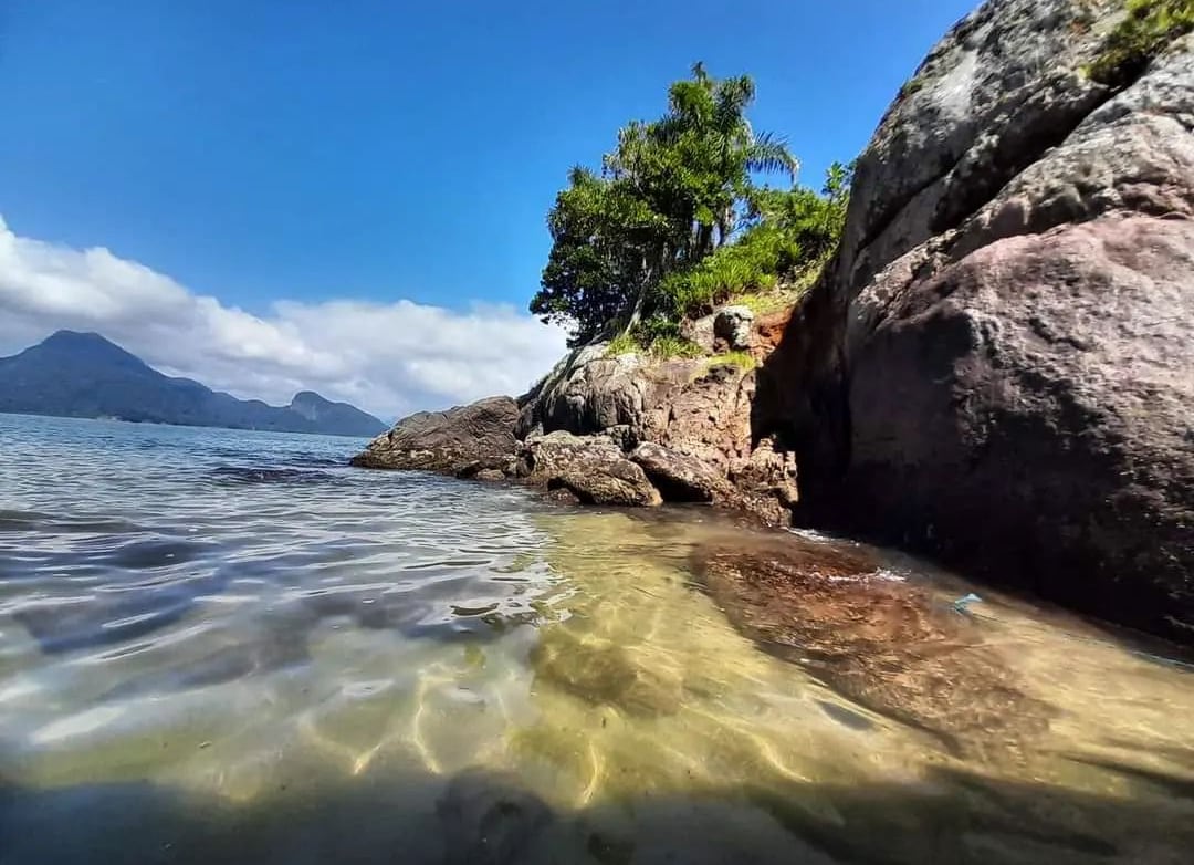 A rocky shoreline is visible, with clear water lapping gently against the stones. Lush greenery is seen on the hillside, alongside distant mountains and a clear blue sky, creating a serene coastal scene.