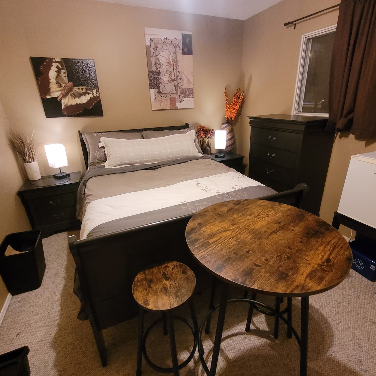 A cozy bedroom features a comfortable bed with neutral bedding and artistic wall decor. Soft lighting from two lamps complements the natural tones. A small, round table with a stool occupies one corner, while a dresser adds to the room’s functionality. A window provides natural light.