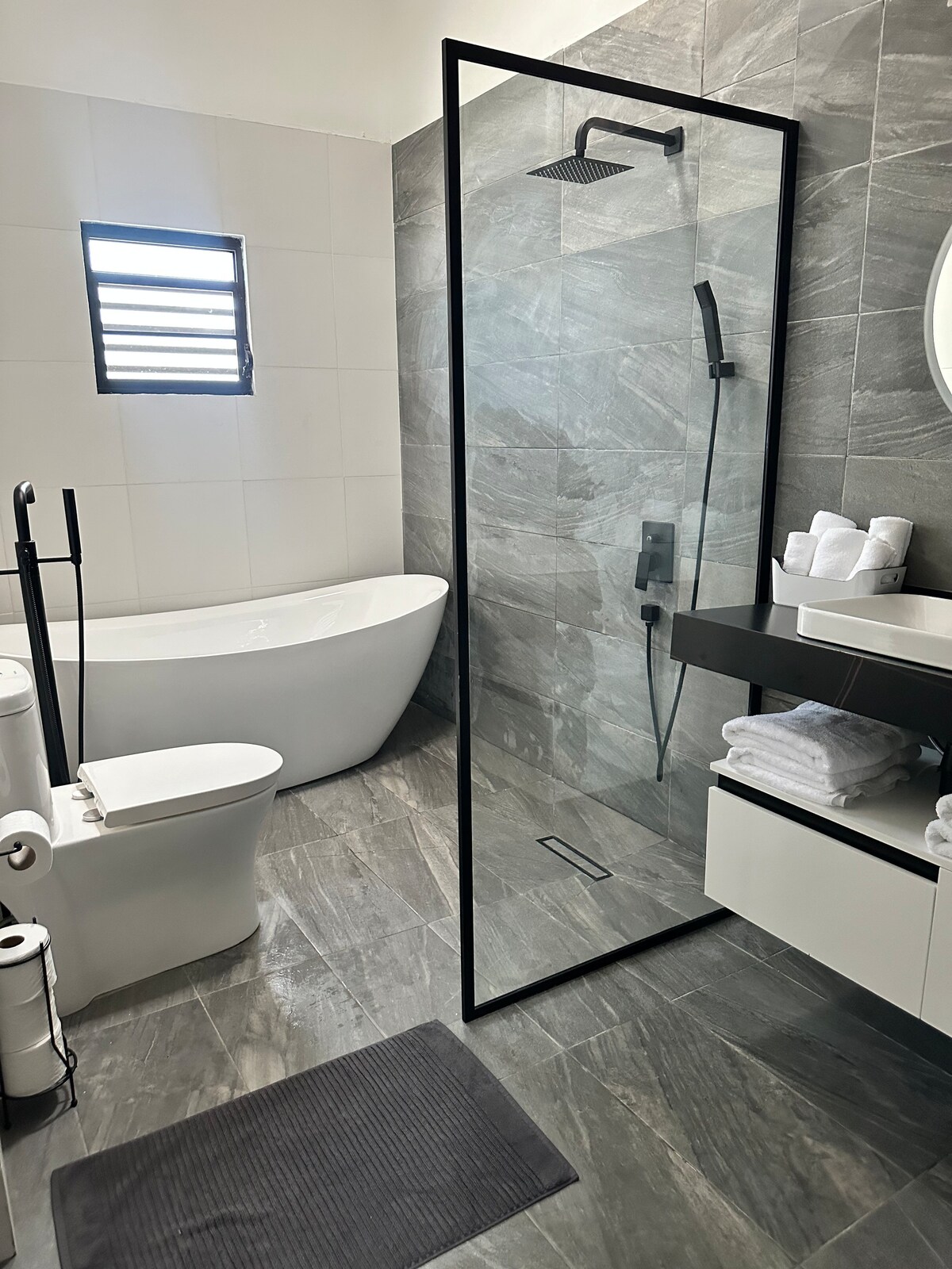 A modern bathroom is featured with a freestanding bathtub and a framed glass shower. Grey tiles provide a sleek backdrop, complemented by a floating vanity with neatly arranged towels. Natural light enters through the small window, enhancing the minimalist design.