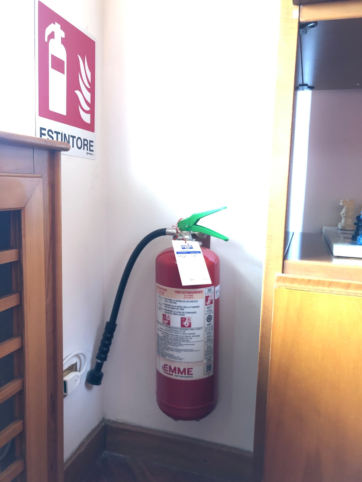 A fire extinguisher is mounted against the wall, accompanied by a safety placard. The extinguisher features a green handle and is securely tagged. Nearby, a wooden cabinet is visible, providing additional context to the room's layout.