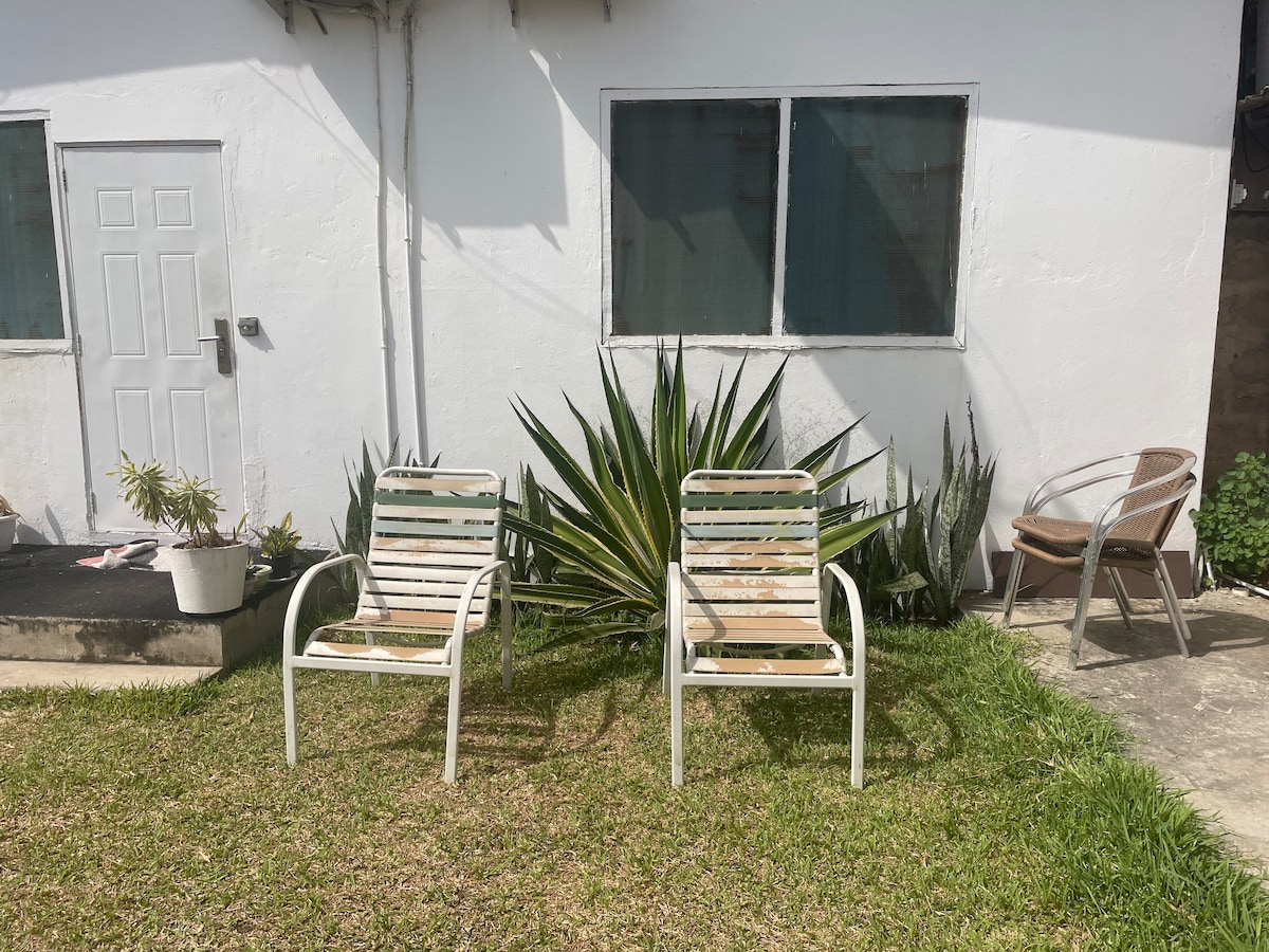 Two striped outdoor chairs are positioned on a grassy lawn, surrounded by plants. A small white pot with a plant is visible nearby. A door and window are seen on the white exterior wall of the building in the background.