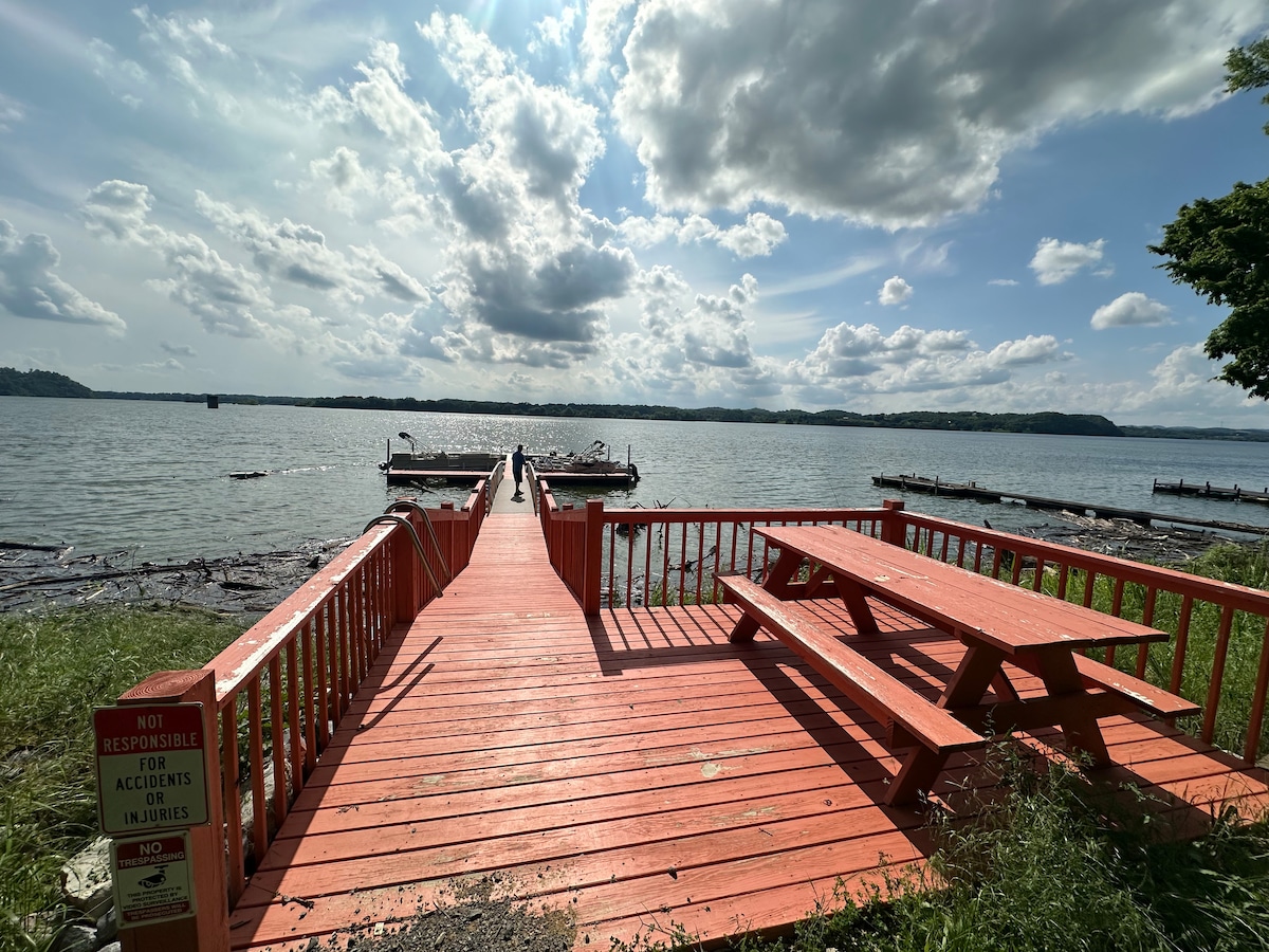 A wooden dock extends into the lake, framed by a bright red railing. A picnic table is positioned on the deck overlooking the water, where boats can be seen in the distance. The sky is partly cloudy, providing natural light that enhances the serene view.