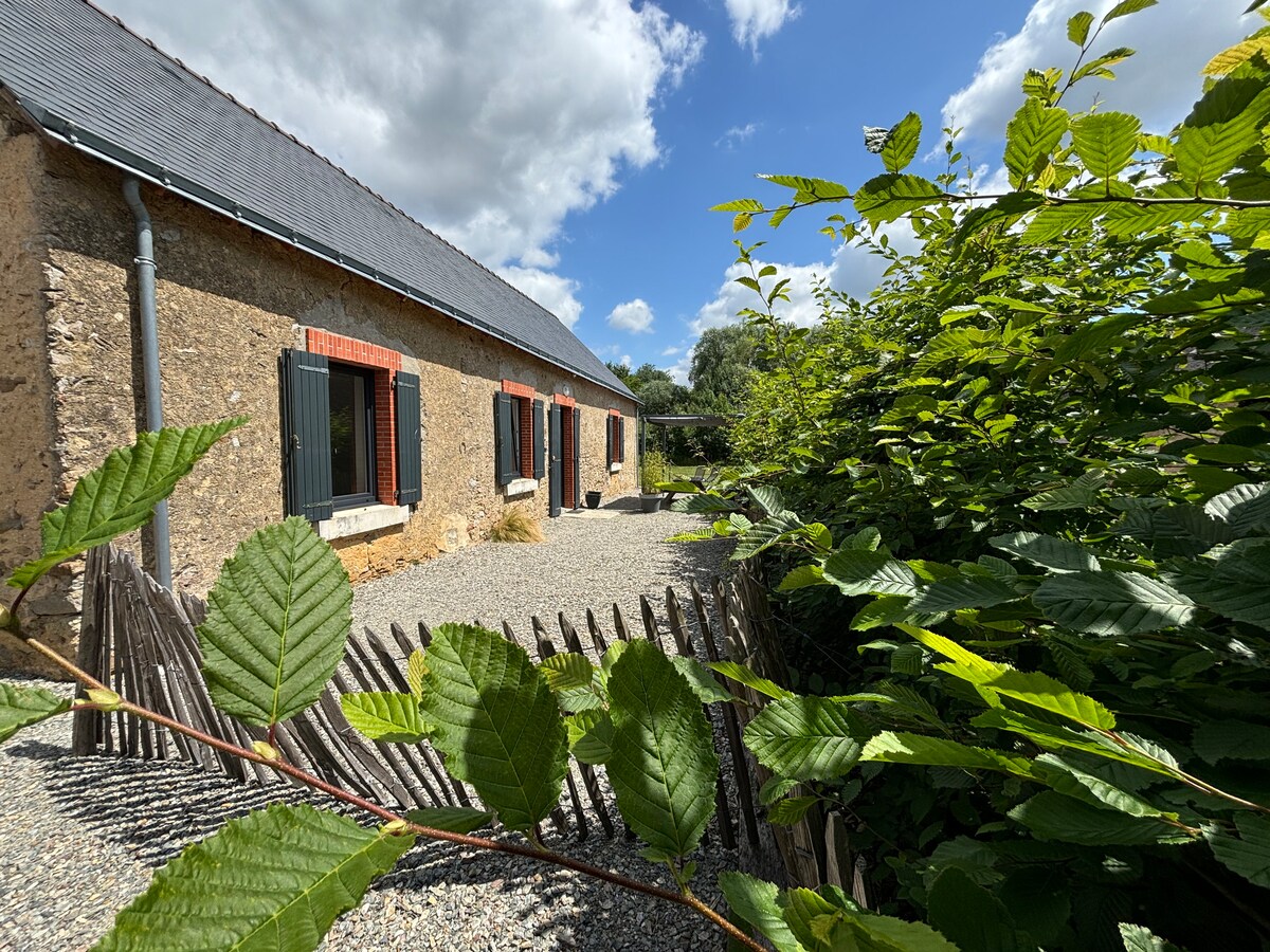 A stone house is framed by lush greenery and a rustic wooden fence. The exterior features multiple windows with dark shutters, offering a glimpse of the gravel path leading to the entrance. A clear blue sky with scattered clouds provides a bright backdrop.