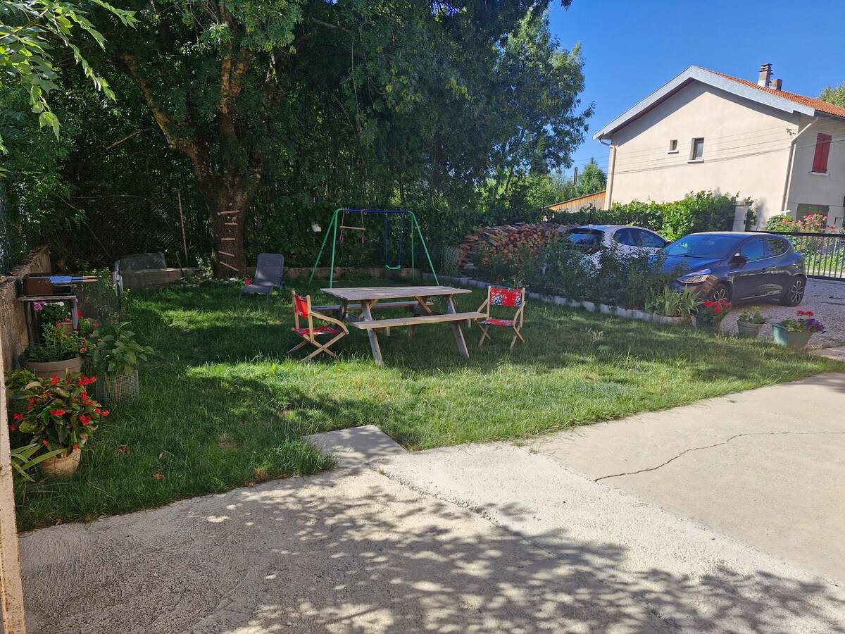 A shared garden space features a grassy area with a wooden picnic table surrounded by eight chairs. Colorful flowers are planted along the edges. In the background, a playground swing set is visible, with a few vehicles parked nearby under the shade of trees.