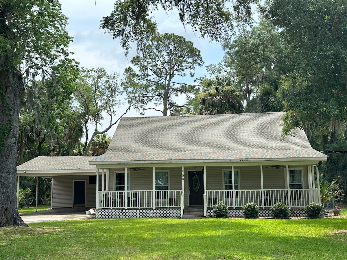 A charming one-story house with a pitched roof is set within a lush green yard. A covered porch with railings invites relaxation, while landscaped shrubs frame the entrance. The carport is adjacent, surrounded by tall trees and greenery, creating a serene outdoor ambiance.