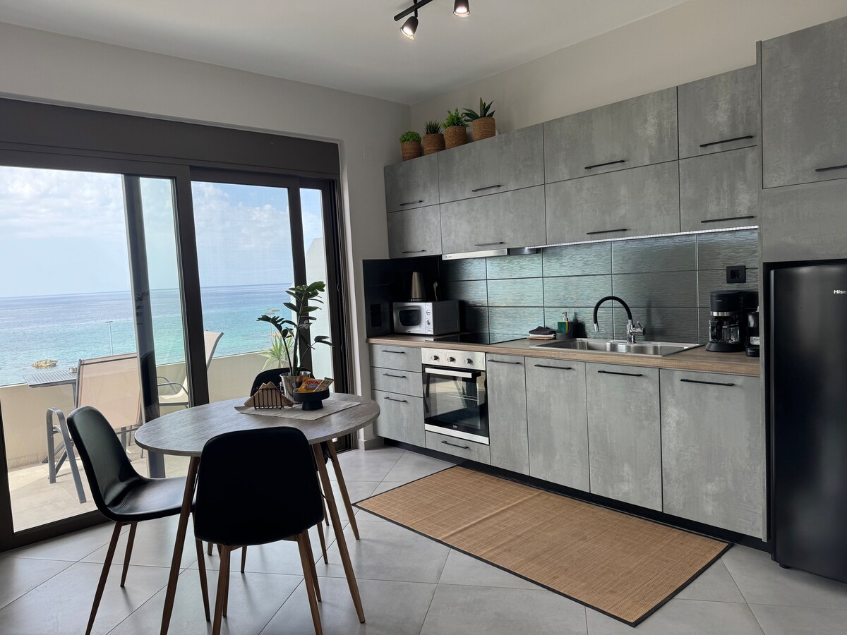 A modern kitchen area features sleek gray cabinetry and countertops. A dining table with black chairs is positioned beside large glass doors that open to a balcony with sea views. Small plants adorn the shelves, enhancing the airy atmosphere of the space.
