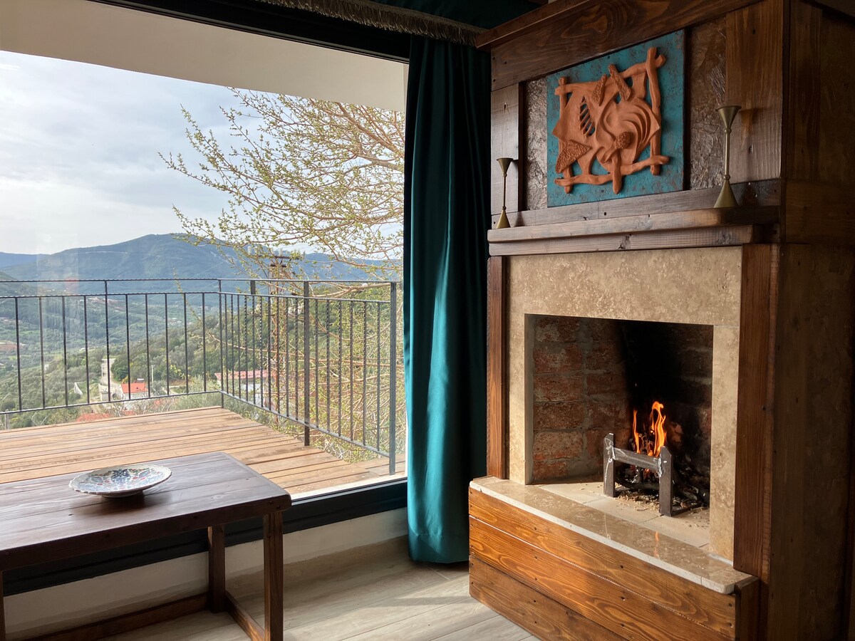 A stone fireplace is positioned beneath a decorative wooden panel, with a fire gently burning. A wooden table sits nearby, while large glass doors frame a scenic mountain view. Light drapes in teal surround the space, creating a cozy ambiance.