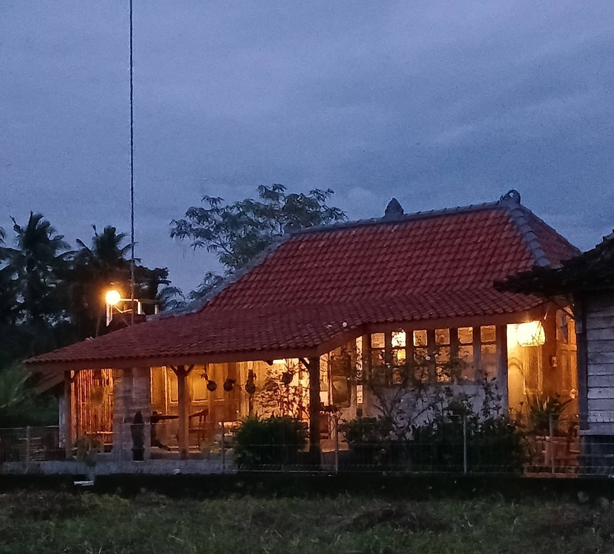 A traditional villa is set amid lush greenery, showcasing a terracotta-tiled roof. Warm light emanates from the windows, hinting at a cozy ambiance inside. The surrounding fields create a tranquil rural setting, with palm trees visible in the background under a softly illuminated evening sky.