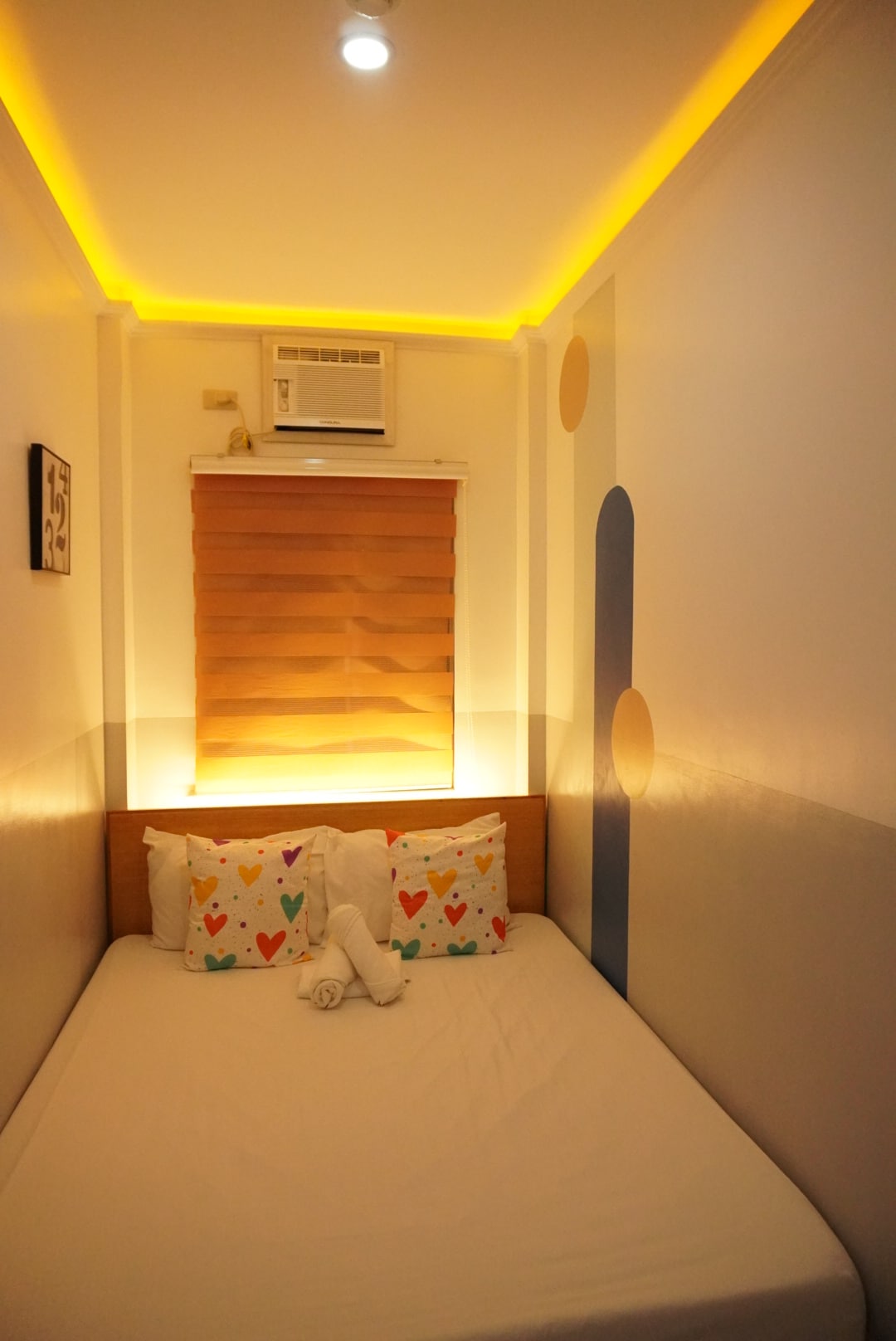 A cozy capsule room features a queen bed adorned with decorative pillows in a heart pattern. Soft ambient lighting from a ceiling fixture enhances the space. A roller blind covers a window, and an air conditioning unit is visible above the bed.