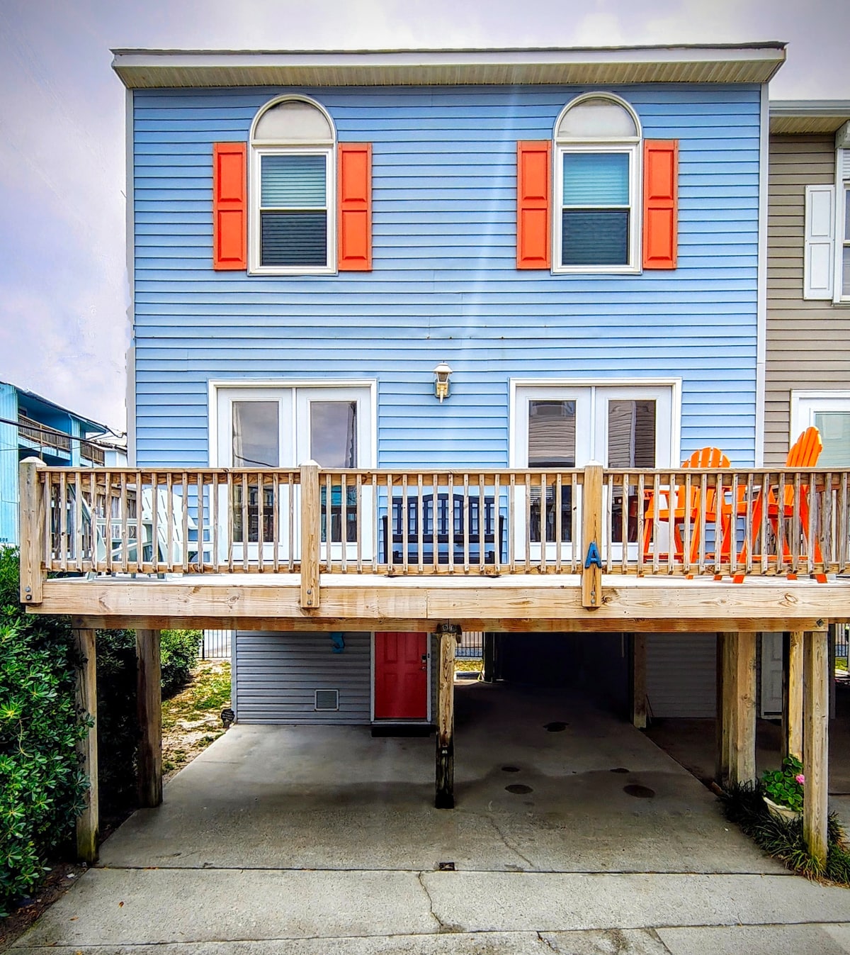 The exterior of a two-story townhome showcases blue siding with bright orange shutters. A large wooden deck with orange captains chairs is visible at the front, while an open parking space is located underneath. The entrance features two large doors, enhancing accessibility.