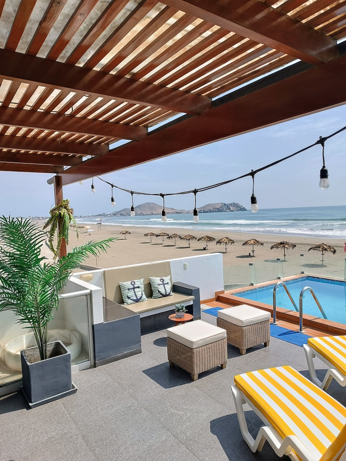 An outdoor terrace features comfortable seating and sun loungers with yellow and white stripes. The view shows the sandy beach and ocean, with umbrellas visible in the distance. Soft lighting is provided by hanging bulbs, contributing to a relaxed atmosphere.