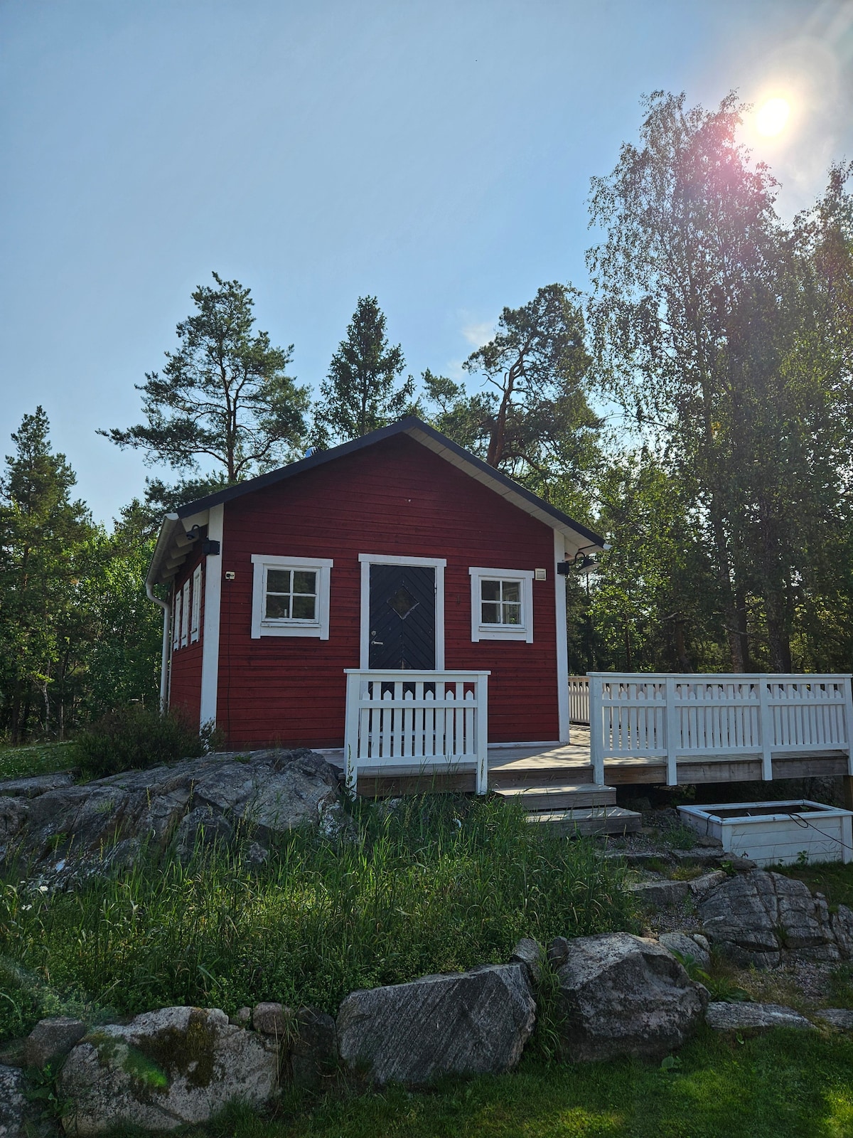 A charming red cottage is positioned on a rocky outcrop, surrounded by lush greenery. A wooden deck leads to the entrance, providing an inviting transition between indoor and outdoor spaces. Sunlight filters through the treetops, creating a tranquil ambiance.