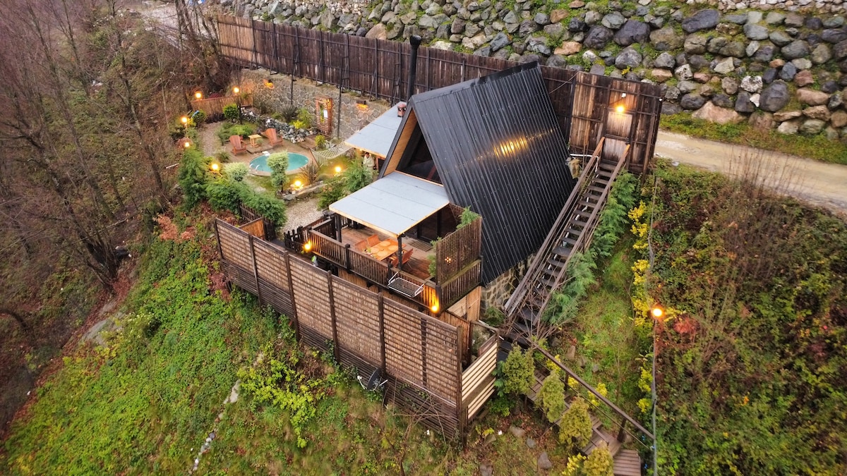 The image displays a secluded, luxurious bungalow surrounded by greenery and positioned against a stone backdrop. Warm lights illuminate the exterior, highlighting a private garden and an inviting hot tub. A pathway leads to the bungalow, enhancing the sense of privacy and tranquility.