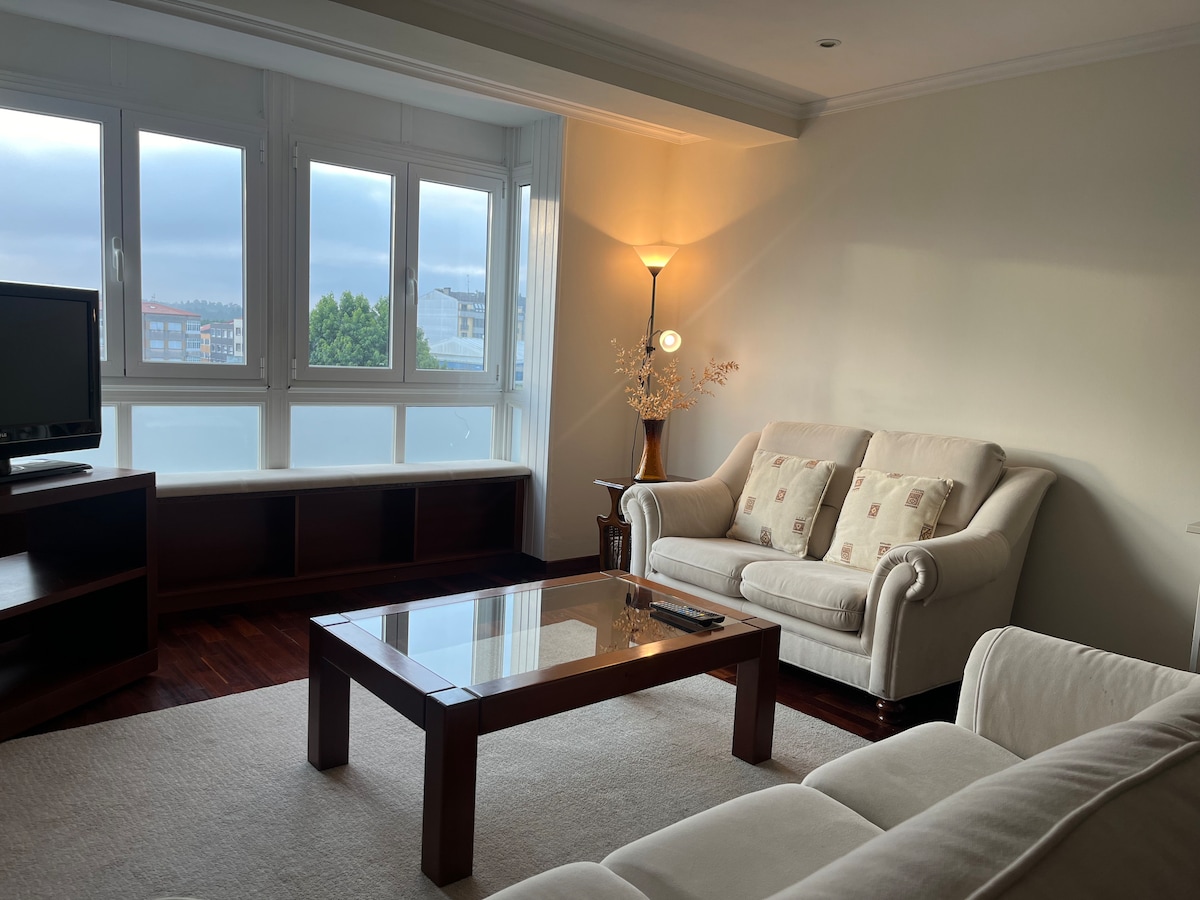 A living area is presented with a light color palette, featuring a comfortable cream sofa and a matching armchair. A wooden coffee table sits in the center, and large windows allow natural light to enter the space, providing views of the surrounding area.