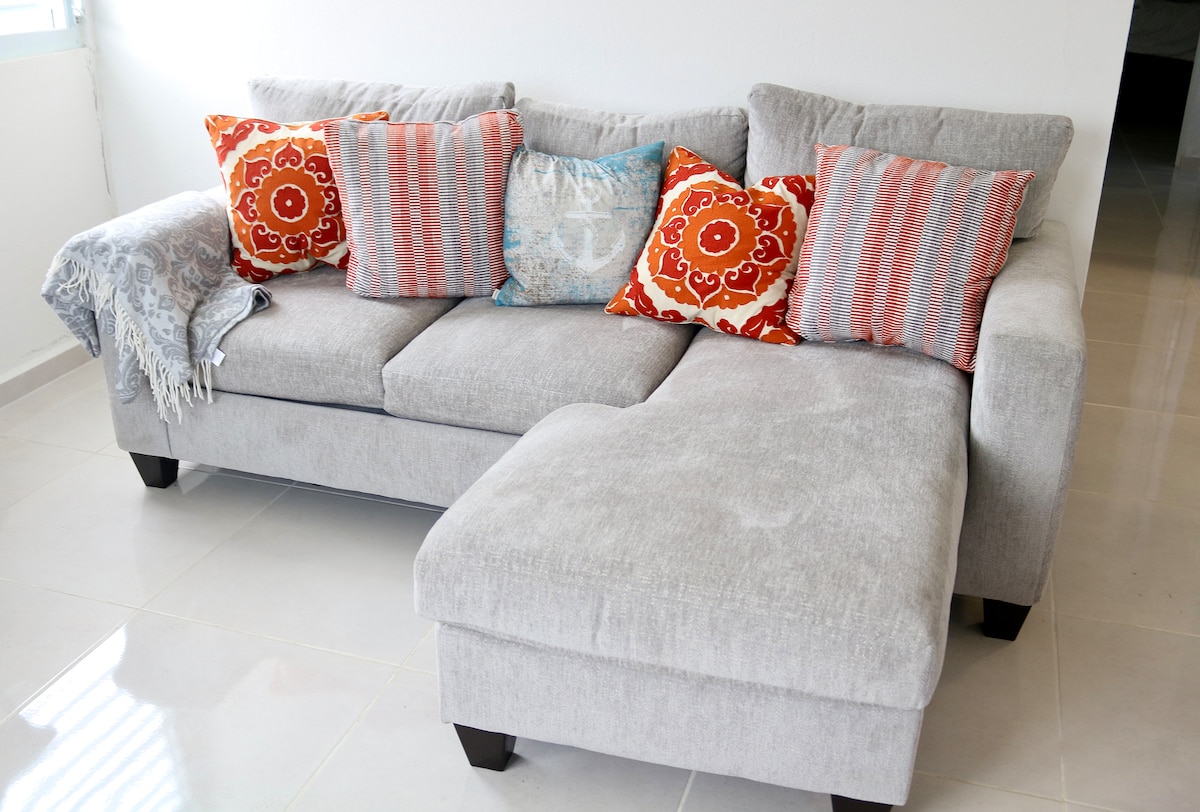 A modern sectional sofa is featured, upholstered in light gray fabric. Multiple decorative cushions in shades of red, orange, and blue are arranged on the seating area. A soft throw blanket is draped over one arm, enhancing the comfortable look of the space.