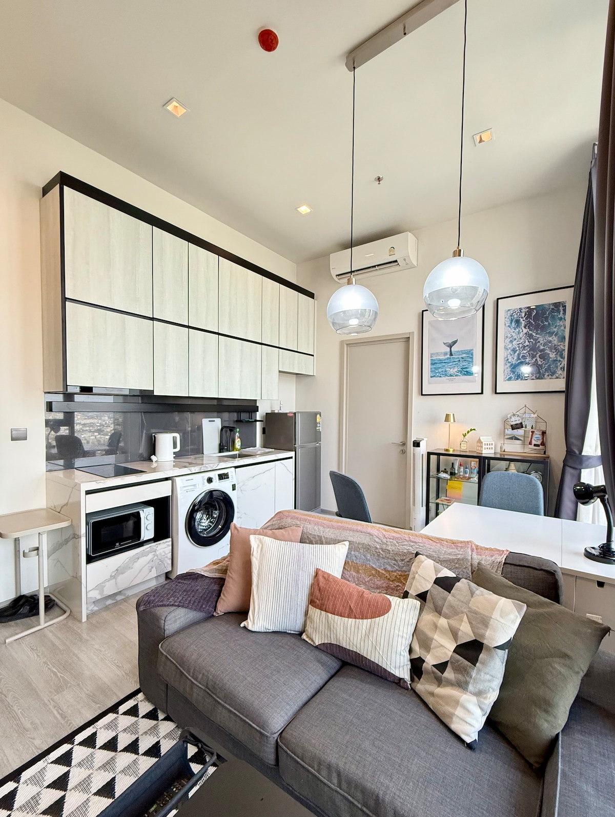 A modern kitchen area features light wood cabinetry with integrated appliances, including a microwave and washing machine. A small dining table with grey chairs is positioned beside a comfortable sofa adorned with decorative cushions. Natural light enters through nearby windows, highlighting the open layout.